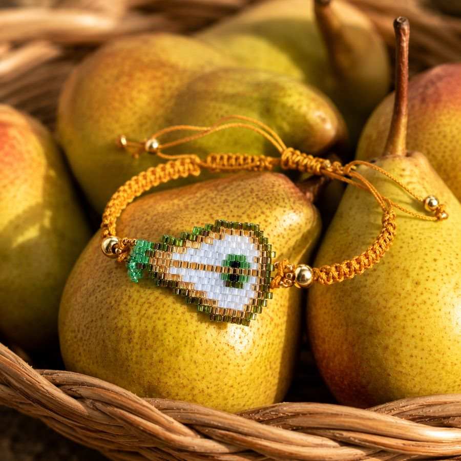 Pulsera Ajustable Pera de Cristal Miyuki: Joyería Artesanal Chic con Macramé Mostaza • Joyas Coquetas pulsera ajustable pera en fondo con cesta de mimbre con peras. Hecha a mano por Joyas Coquetas en Málaga