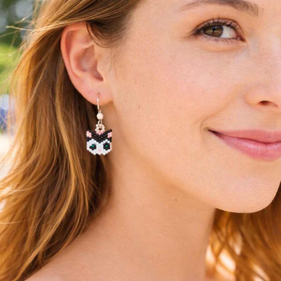 Pendientes Gata Artesanales • Joyas Coquetas Primer plano de una joven sonriente que lleva unos pendientes de gato hechos a mano con cuentas Miyuki blancas y negras. Suave iluminación natural exterior. Pendientes hechos a mano por Joyas Coquetas en España.