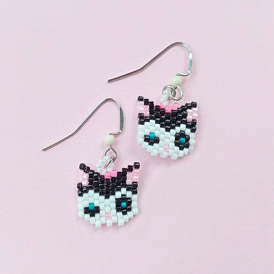 Pendientes Gata Artesanales • Joyas Coquetas Bonitos pendientes de gatitos blancos y negros hechos a mano con cuentas de cristal, colocados sobre un fondo rosa pastel suave para crear un efecto coqueto. Hechos por Joyas Coquetas en España.