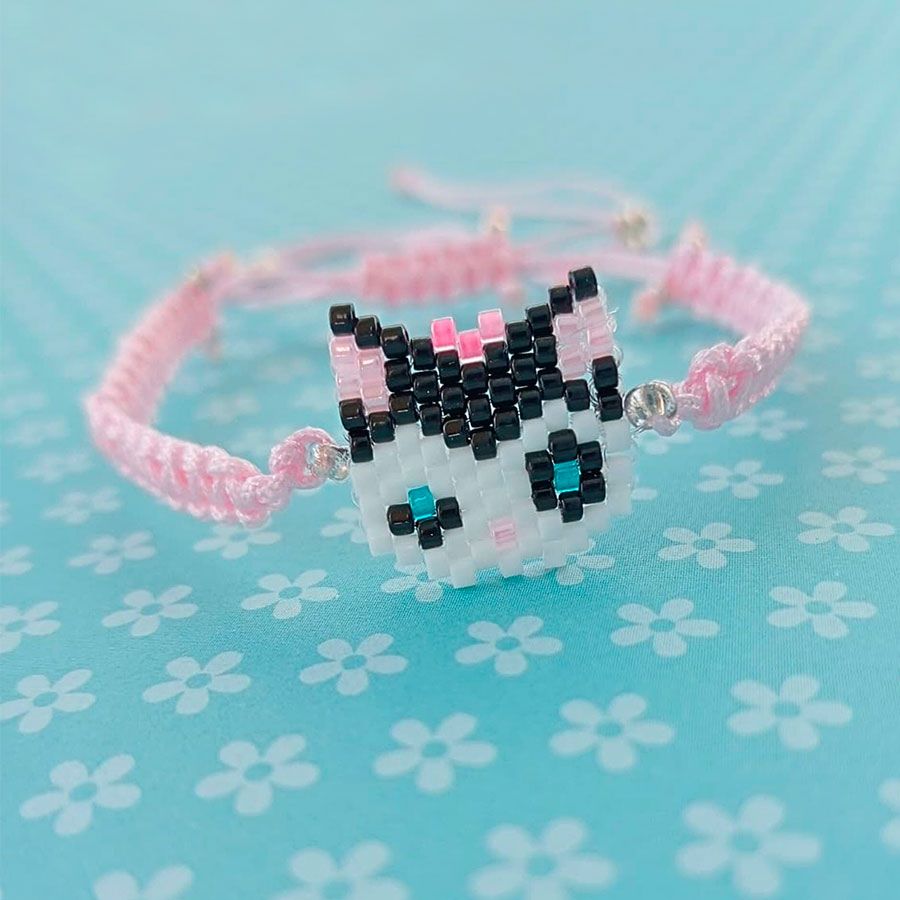 Pulsera Gatita Lazo Rosa • Joyas Coquetas Pulsera Gatita Lazo Rosa con charm de gatita tejido en cristal Miyuki sobre fondo azul floral. Hecha a mano por Joyas Coquetas en España.