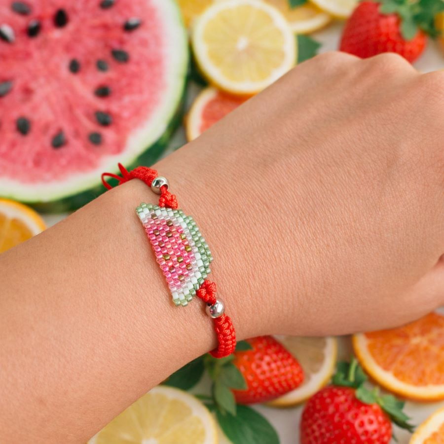 Pulsera sandía ajustable • Joyas Coquetas Pulsera sandía artesanal puesta en la muñeca con sandía y fresas de fondo.