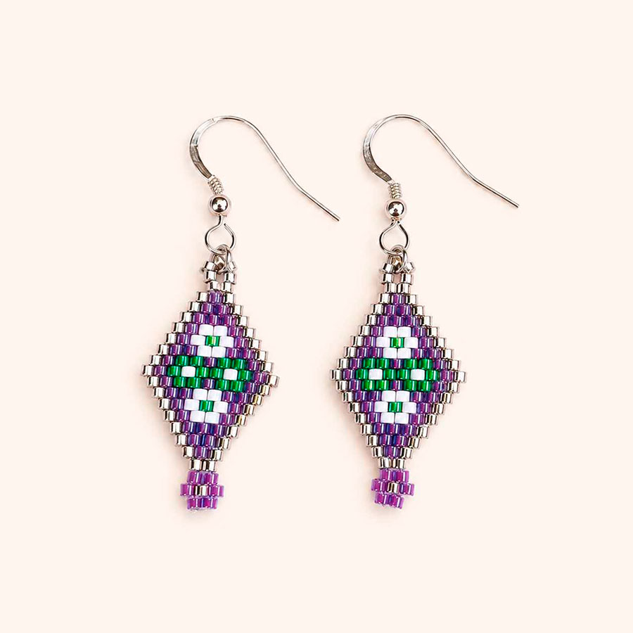 Pendientes florales Miyuki artesanales en tonos morado, verde y plata sobre fondo beige claro. Hechos a mano por Joyas Coquetas en Málaga.